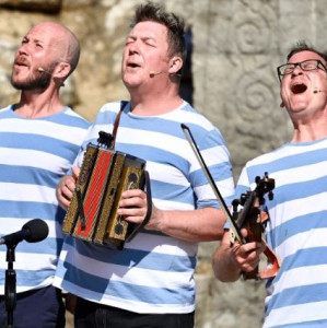 A photo of three men singing, all of them are wearing blue and white stripe tops. One is playing an accordion.