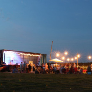 A photograph of lots of people sitting in a field at night under festoon lighting.  They are sat next to a lorry that is also a music stage.  There is a lot of night sky in the photograph and it says 'Chill to a Tune in June'. 