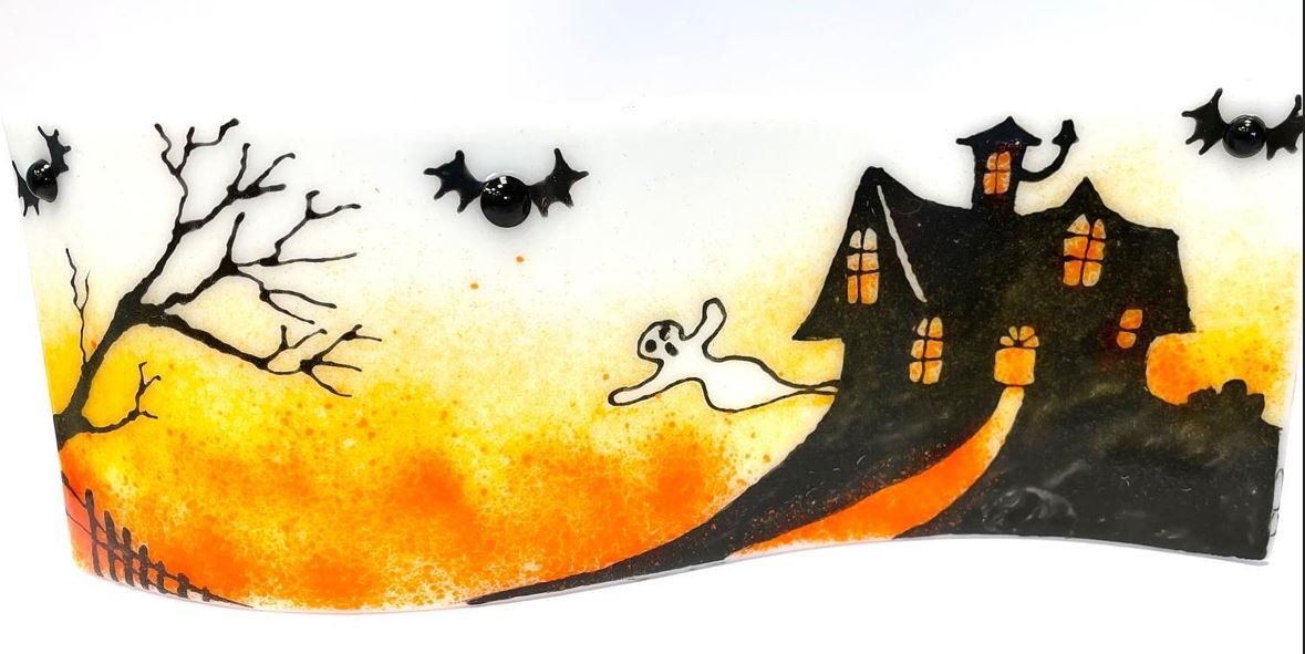 A photograph of a fused glass Halloween scene. It includes a haunted house and bats in black silhouetted on a white and orange coloured glass. There is also a white ghost.